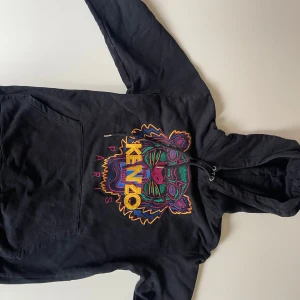 Svart Kenzo hoodie med tigerbrodyr - Svart hoodie från Kenzo med ikonisk färgglad tigerbrodyr och Kenzo Paris-text på bröstet. Tröjan har huva med snörning och en stor magficka. Perfekt statement-plagg med cool streetkänsla och snygga detaljer i flera färger.
