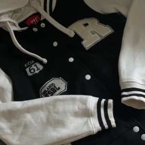 Svart och vit bomberjacka med huva - Snygg svart och vit bomberjacka med huva och coola patches på bröstet. Jackan har vita ärmar, svarta och vita muddar samt knappar framtill. Stora bokstaven R och broderade detaljer ger en sportig collegekänsla. Perfekt för dig som gillar streetstyle.