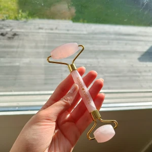 Rosenkvarts ansiktsroller - Säljer en lyxig ansiktsroller i äkta rosenkvarts med guldfärgade detaljer. Perfekt för att boosta din hudvårdsrutin och ge ansiktet en fräsch känsla. Handtaget är rosa och har texten 'ROSENKVARTS' i guld. Enkel att använda och snygg att ha framme.