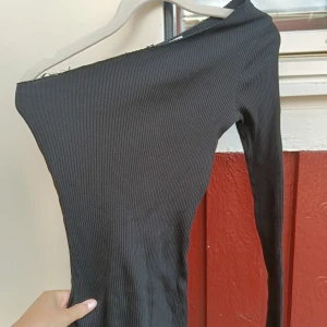 Svart oneshoulder ribbad topp - Snygg svart ribbad toppmed oneshoulder-design och lång ärm. Klänningen är figurnära och har en cool asymmetrisk halsringning som ger en trendig vibe. Perfekt för dig som gillar minimalistisk stil med en twist.