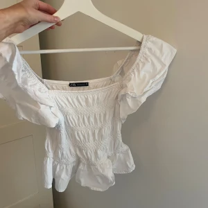Vit offshoulder topp från Zara - Supersöt vit offshoulder topp från Zara med smockad midja och volang nertill. Toppen har korta puffärmar och är tillverkad i mjuk bomull. Perfekt för en fräsch och somrig look!