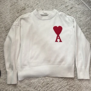 Vit stickad tröja från Ami Paris - Vit stickad tröja från Ami Paris med röd hjärtlogga och bokstaven A på bröstet. Tröjan har ribbade muddar och krage, samt lång ärm. Perfekt för dig som gillar stilrena plagg med en twist.