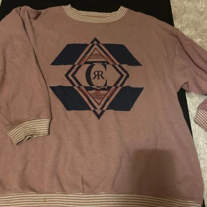 Rosa sweatshirt från Cerruti 1881 - Säljer en rosa sweatshirt från Cerruti 1881 med coolt grafiskt tryck i mörkblått framtill. Tröjan har långärmade ärmar och ribbade muddar med vita och rosa ränder vid hals, ärmslut och nederkant. Perfekt för en avslappnad stil.