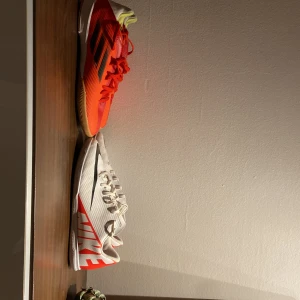 Nike och Adidas fotbollsskor inomhus - Säljer två par inneskor för alla sporter. Två par snygga inomhusfotbollsskor: ett par vita Nike med svart swoosh och röd logga, samt ett par Adidas i orange och rött med svarta ränder. Båda har snörning, platt sula och är gjorda i syntetmaterial. Perfekta för snabba matcher på inomhusplan! Kan förhandlas med priset vill bara få dem sålda