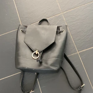 Svart ryggsäck med guldring - Snygg svart ryggsäck i skinnimitation med justerbara axelband och lock med dekorativ guldfärgad ring. Praktisk och stilren modell med dragsko under locket, perfekt för att bära dina grejer till skolan eller stan.
