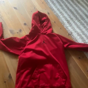 Röd Jordan hoodie med huva - Snygg röd hoodie från Jordan med klassisk svart Jumpman-logga på bröstet. Tröjan har huva och en stor magficka framtill. Tillverkad i mjukt material som känns skönt mot huden. Perfekt för dig som gillar sportig och avslappnad stil.