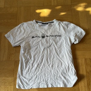 Vit t-shirt från Sail Racing - Vit t-shirt från Sail Racing med svart logga tryckt framtill. Klassisk rund halsringning och korta ärmar. 