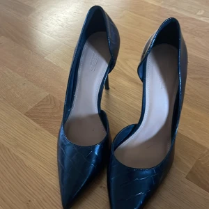 Svarta pumps med spetsig tå - Snygga svarta pumps med spetsig tå och hög klack. Skorna har en glansig finish med krokopräglat mönster som ger en lyxig känsla. Perfekta för dig som vill sticka ut med en elegant och trendig look.