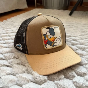 Capslab keps med Kalle Anka Disney - Snygg keps från Capslab x Disney med Kalle Anka-motiv framtill. Framsidan är beige och brun, baksidan är svart mesh för extra ventilation. Broderad detalj på sidan och justerbar passform.