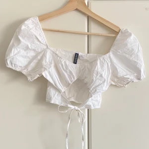Vit croppad blus från Divided - Supersöt vit croppad blus från Divided med puffärmar och knytning i midjan. Toppen har fyrkantig urringning och är tillverkad i tunn bomull, perfekt för varma dagar. Den har en feminin och trendig look med voluminösa ärmar och öppen detalj framtill.