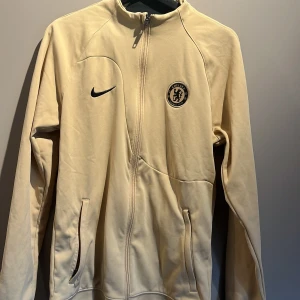 Chelsea x Nike beige track jacket S - Chelsea FC track jacket från Nike i beige med svart logga och klubbmärke på bröstet. Slim fit-modell med hel dragkedja framtill och två fickor. Perfekt för dig som vill representera ditt lag med stil. Köpt på Chelseas officella klubb shop helt oanvänd.