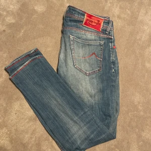 Jacob Cohen jeans - Snygga blåa jeans från Jacob Cohen med coola röda sömmar och knappar. Skick 7,5/10 då läderlappen där bak e sönder. Priset är ej hugget i sten