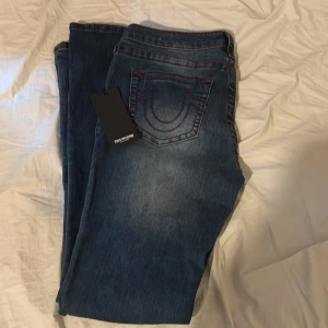 True Religion blå jeans med röd söm - Säljer ett par snygga blå jeans från True Religion med ikoniska röda kontrastsömmar och tydliga bakfickor med broderad logga. Klassisk femficksmodell och silverfärgade knappar. Perfekt för dig som vill sticka ut med coola detaljer och streetkänsla. Aldrig gått ut med dessa bara provat på. Pris kan diskuteras💕