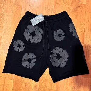 Svarta shorts från Denim Tears med mönster - Svarta shorts från Denim Tears med unikt grått blommigt mönster över hela plagget. De har elastisk midja med snörning för skön passform. Perfekta för dig som vill sticka ut med streetwear-vibe och cool design.