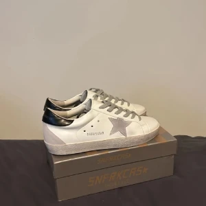 Vita sneakers med grå stjärna - Snygga vita sneakers från GGDB/SSTAR med grå mockadetaljer och en stor grå stjärna på sidan. Skorna har grå snörning, svart hälparti och platt sula. Tillverkade i skinn och mocka för en clean och trendig look.