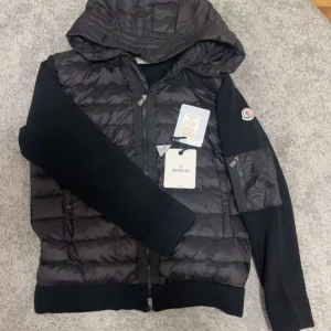 Svart Moncler pufferjacka med huva - Svart pufferjacka från Moncler med stickade ärmar och vadderad kropp. Jackan har huva, dragkedja framtill och en extra ficka med dragkedja på ärmen. Klassisk Moncler-logga på vänster ärm. Perfekt för dig som vill ha en snygg och sportig look.