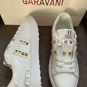 Valentino Rockstud vita sneakers - Snygga vita Valentino Rockstud sneakers i skinn med ikoniska guldiga nitar och chunky platt sula. Klassisk rund tå, snörning och coola detaljer längs sidorna och tungan. Perfekta för dig som vill sticka ut med lyxig streetstyle.