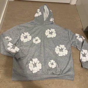 Grå hoodie med vita blommor - Säljer en grå hoodie med stora vita blommor tryckta över hela tröjan. Hoodien har en klassisk huva och känguruficka framtill. Perfekt för dig som gillar en chill och unik stil. Materialet känns mjukt och skönt mot huden.