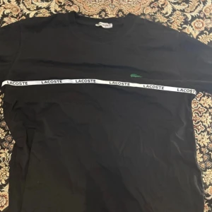 Svart Lacoste t-shirt med logoband - Svart t-shirt från Lacoste i bomull, med klassisk rund hals och korta ärmar. Snyggt vitt band med Lacoste-tryck går horisontellt över bröstet och den ikoniska gröna krokodilen är broderad på vänster sida. Perfekt för en clean och sportig look.