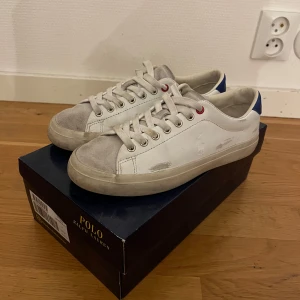 Vita sneakers från Polo Ralph Lauren - Snygga vita sneakers från Polo Ralph Lauren med grå mockadetaljer på tån och blå häl. Väldigt unika skor med en best down look, det påminner och liknar lite som de populära skorna golden goose och Philippe model. Skorna passar väldigt bra till en bra look i vardagen. Nypris: 1300kr. Skorna är lite smutsiga med det går att tvätta lätt. Hör av er vid minsta lilla funderingar. 