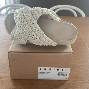 Beige flätade sandaler från Inuikii - Snygga sandaler från Inuikii i beige med breda, flätade band i textil som korsar över foten. Platt sula och öppen tå ger en chill vibe, perfekt för varma dagar. Stilren design som sticker ut med sitt grova, stickade material.
