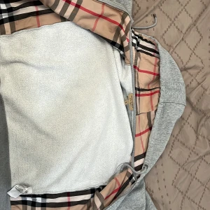Grå hoodie från Burberry med rutor - Säljer en grå hoodie från Burberry i storlek M men koftan sitter som en S . Utsidan är ljusgrå med dragkedja och huva, insidan har det klassiska Burberry-rutiga fodret i beige, svart och rött. Mjuk och skön, perfekt för dig som vill ha en stilren men ändå lyxig look. Pris kan diskuteras 