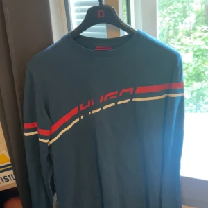 Blå långärmad Hugo tröja - Snygg blå långärmad tröja från Hugo med röda och vita ränder över bröstet och armarna. Klassisk crewneck och tryckt logga framtill. Perfekt för dig som gillar sportig och stilren look.