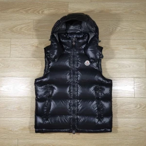 Svart dunväst från Moncler med huva - Snygg svart dunväst från Moncler med avtagbar huva och två dragkedjeförsedda fickor. Västen har en glansig finish och klassisk Moncler-logga på bröstet. Perfekt för lager-på-lager och streetstyle.