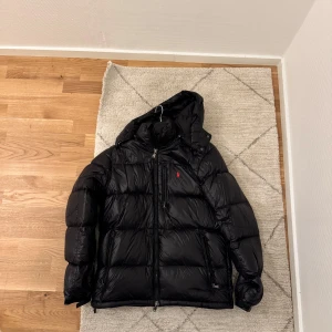 Polo Ralph Lauren vinterjacka - Säljer en svart dunjacka från Polo Ralph Lauren med huva och dragkedja. Jackan har en klassisk pufferstil, röda loggan broderad på bröstet och flera praktiska fickor med dragkedja. Perfekt för kalla dagar och riktigt snygg till streetwear.
