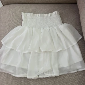 Vit volangkjol med resårmidja - Supersöt vit kortkjol med bred smockresår i midjan och dubbla lager volanger. Kjolen är luftig och lätt, perfekt för varma dagar. Materialet känns som tunn polyester eller liknande, vilket ger ett flowy och romantiskt intryck.