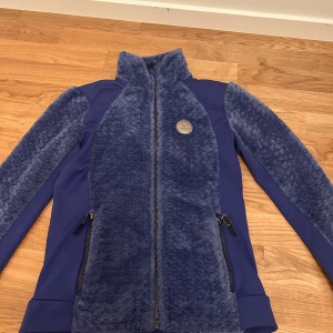 Blå kofta från Jh Collection - Säljer en snygg blå teddyjacka från JHCollection med fluffigt teddytyg på framsidan och ärmarna. Jackan har hel dragkedja, två sidofickor med dragkedja och en cool logodetalj i silver på bröstet. Perfekt för dig som gillar mjuka och trendiga jackor. Passar även perfekt till schabraket jag säljer. Skulle säga att den motsvarar storlek s