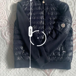 Svart Moncler pufferjacka med huva - Svart pufferjacka från Moncler med quiltad front, huva och dragkedja. Jackan har Moncler-logga på ärmen, ribbade muddar och fickor med dragkedja. Materialmix med glansig polyester framtill och mjuka ärmar. Perfekt för dig som vill ha en stilren och sportig look.