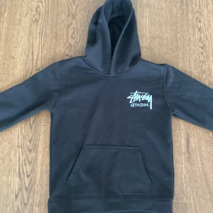 Svart Stüssy hoodie med logga - Svart hoodie från Stüssy med stor vit logga på ryggen och mindre logga på bröstet. Klassisk känguruficka framtill och huva. Skön passform och materialet är mjuk bomullsmix som känns riktigt nice.