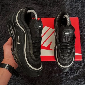 Nike Air Max 97 svarta sneakers - Svarta Nike Air Max 97 sneakers med vita detaljer och ikonisk vågig design. Skorna har synlig Air-sula, snörning och reflekterande ränder. Ovandelen är i mesh och syntet, vilket ger en sportig och modern look. Perfekta för dig som gillar streetstyle och komfort.