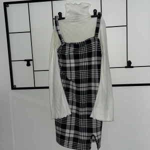 Rutig svartvit kortklänning - Säljer en snygg svartvit rutig kortklänning med breda axelband och liten slits framtill. Klänningen har en cool 90-tals vibe och passar perfekt över en långärmad topp. Materialet känns mjukt och bekvämt, och den rutiga designen gör den extra trendig.