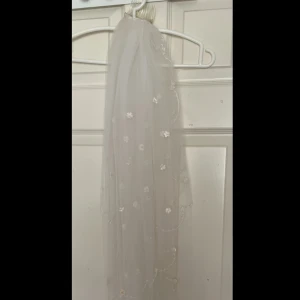 Vit brudslöja med blommor - Säljer en vit slöja med diademfäste, dekorerad med små blommor och pärlor. Slöjan är gjord i tunn, transparent syntet och har en romantisk känsla med broderade detaljer längs kanten. 