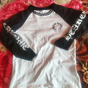 Chrome Hearts svartvit långärmad tröja - Säljer en svartvit långärmad tröja från Chrome Hearts med kontrastfärgade ärmar och stora tryck på både ärmar och rygg. Tröjan har klassisk rund hals och coolt Chrome Hearts-tryck runt halsen och på bröstet. Perfekt för dig som gillar streetwear och statement-plagg.