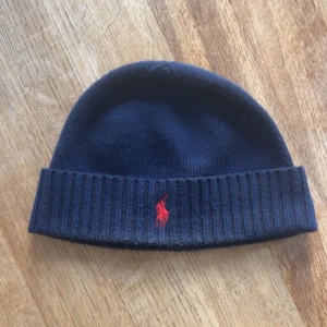 Mörkblå mössa från Polo Ralph Lauren - Snygg mörkblå mössa från Polo Ralph Lauren i mjuk och tvättbar merinoull. Klassisk ribbad kant och ikonisk röd broderad logga framtill. Perfekt accessoar för kyliga dagar och enkel att matcha med olika outfits.