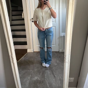 Krämvit blus med pärlknapp - Säljer en stilren krämvit blus med korta ärmar och rund halsringning. Blusen har en snygg pärlknapp framtill som detalj och är tillverkad i ett slätt, lite glansigt material. Perfekt att matcha med jeans eller kjol för en clean look. Använd en gång