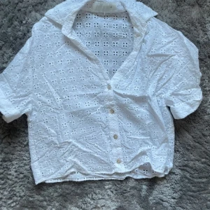 Shorta Zara🤍 - Superfin vit skjorta med korta ärmar och broderade blommor i tyget. Skjortan har knappar framtill och en luftig, croppad passform. Perfekt för varma dagar och ger en fräsch och somrig vibe.