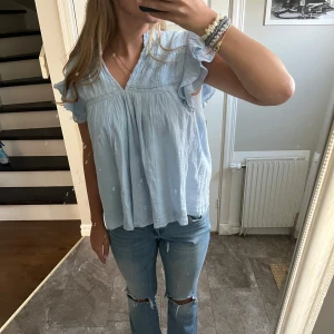 Ljusblå blus med volangärm - Supersöt ljusblå blus med v-ringning och volangärmar. Blusen har fina spetsdetaljer och ett luftigt, flowy fall. Perfekt för dig som gillar en romantisk och avslappnad stil. Materialet känns lätt och svalt, perfekt för varmare dagar.