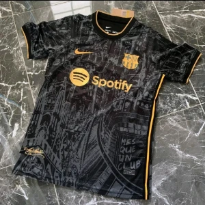 Svart Barcelona Nike Dri-FIT tröja S - Säljer en svart Barcelona fotbollströja från Nike med guldfärgade detaljer och tryck. Tröjan har korta ärmar, Dri-FIT-material och ett unikt stadsmönster över hela tyget. FCB-emblem, Spotify-logga och UNHCR-tryck i gult. Perfekt för dig som älskar Barca!