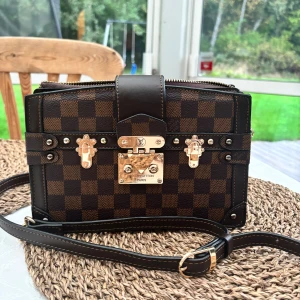 Rutig brun axelväska med gulddetaljer - Säljer en snygg axelväska från i brunt och svart rutigt mönster. Väskan har guldfärgade metalldetaljer, lås och dragkedja samt en justerbar axelrem. Perfekt för dig som gillar lyxiga accessoarer med attityd. Köpt fast har inte kommit till användning 