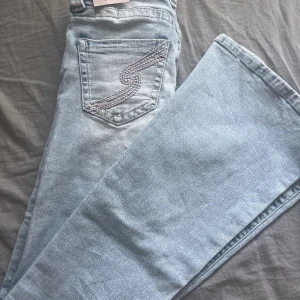 Ljusblå low waist jeans från Gina Tricot - Säljer ett par ljusblå jeans från Gina Tricot i modellen dixie ultra low jeans. Byxorna har låg midja, raka ben och snygga broderade detaljer på bakfickorna. Klassisk femficksmodell i mjukt jeanstyg, perfekta till en avslappnad look.