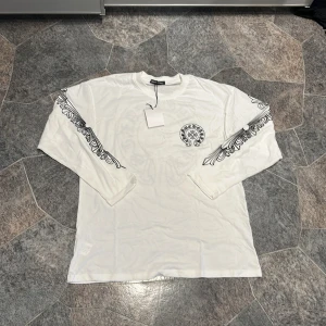 Vit långärmad Chrome Hearts tröja - Vit långärmad tröja från Chrome Hearts med svart tryck på bröstet och ryggen samt detaljerade svarta mönster längs ärmarna. Tröjan har en klassisk passform och är gjord i mjuk bomull. Perfekt för dig som gillar streetwear med unika detaljer.