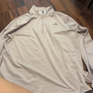 Ljuslila half zip tröja från Adidas - Säljer en ljuslila långärmad tröja från Adidas med half zip och hög krage. Perfekt för träning eller chill dagar. Tillverkad i mjukt och lätt polyestermaterial som andas. Adidas-logga på bröstet ger en sportig vibe.