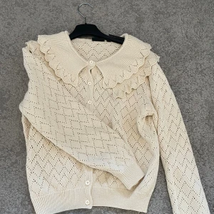Beige kofta med volangkrage från KappAhl - Supersöt beige kofta från KappAhl i stickad bomull med genomgående knappar och långärmad modell. Koftan har ett hålmönster i diamantform och en bred volangkrage som ger en gullig vintagekänsla. Perfekt att styla med jeans eller kjol!