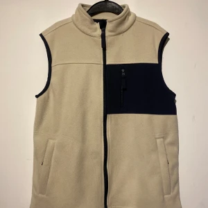Beige fleeceväst från A Day's March - Snygg beige fleeceväst från A Day's March med marinblå detaljer och dragkedja framtill. Västen har hög krage, två sidofickor och en bröstficka med dragkedja. Perfekt att slänga på över en hoodie för extra värme och stil. Nypris på denna fantastiska väst är 1200 kr men köp den för endast 499 kr hos oss😀