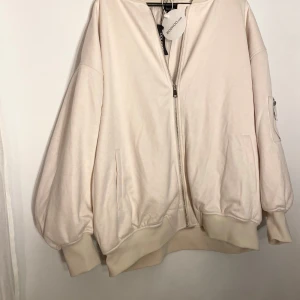 Oversized beige bomberjacka från Boohoo - Oversized beige bomber jacka från Boohoo med dragkedja framtill, breda muddar och fickor på sidorna. Extra ficka med dragkedja på ena ärmen. Fusk mocka och avslappnad passform, perfekt för lager-på-lager. Snygg och enkel att styla.