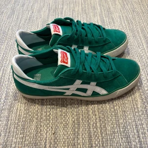 Gröna Onitsuka Tiger sneakers - Snygga gröna Onitsuka Tiger sneakers med vita detaljer och klassisk logga på plösen. Skorna har snörning, vit sula och är tillverkade i mocka och syntet. Perfekta för dig som gillar retrostil och vill sticka ut med färg på fötterna.